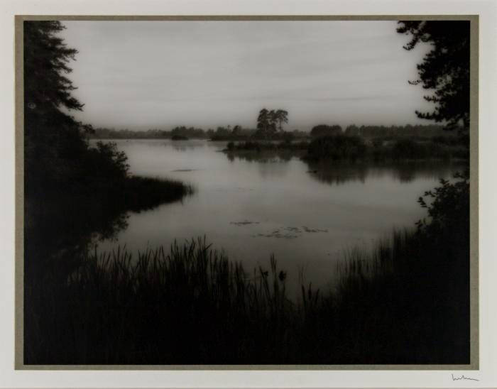 Black and white photograph of a small body of water surrounded by grass and trees. Area looks misty.